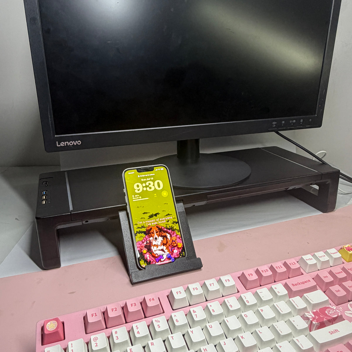 Command Monitor Riser under a monitor with its phone stand extended, holding a phone upright