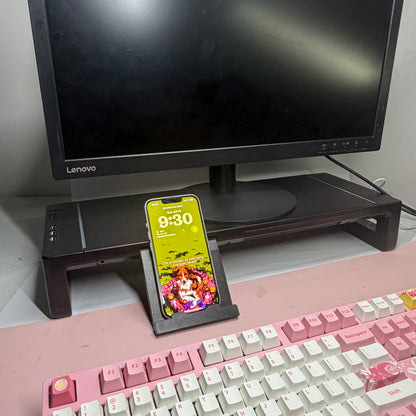 Command Monitor Riser under a monitor with its phone stand extended, holding a phone upright
